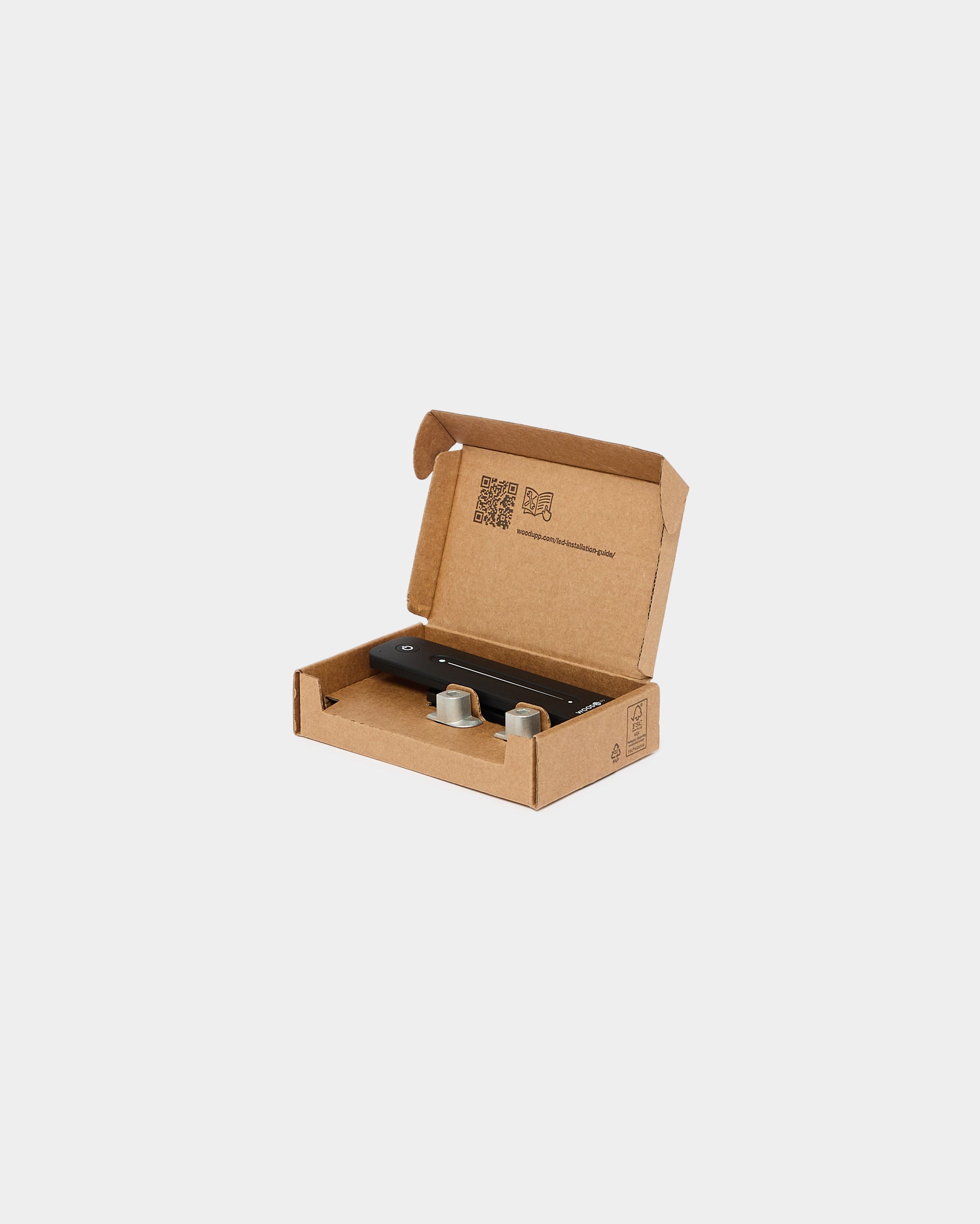 Open brown cardboard box containing two small metallic accessories and a black rectangular mounting piece, suitable for assembling acoustic panel products, on white background.